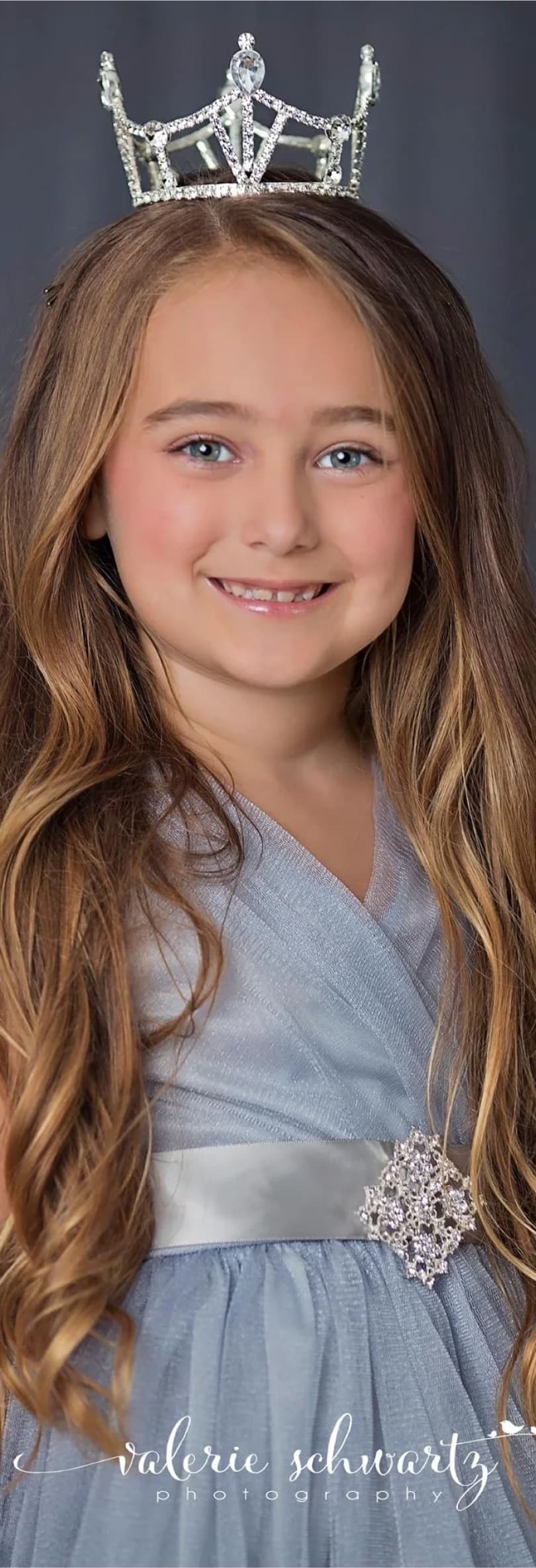 Smiling little girl with a crown on her head dressed in an elegant blue-gray outfit with a rhinestone sash